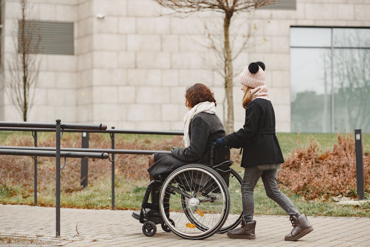 About A young girl helps a woman in a wheelchair on a sidewalk, showing care and support.